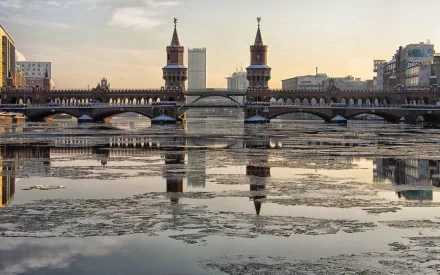 HD PC desktop wallpaper: Berlin's Oberbaum Bridge, a twin-towered man-made bridge over the Spree, reflected in partially frozen water at sunrise.