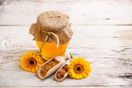A jar of honey wrapped in burlap sits on a wooden table alongside sunflower flowers and wooden scoops filled with pollen, creating a vibrant and natural food-themed backdrop.