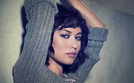 HD PC desktop wallpaper featuring celebrity Olga Kurylenko in a cozy gray sweater, posing with arms raised against a muted background.