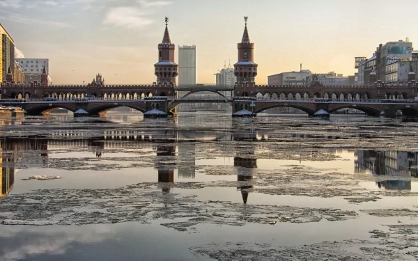 HD PC desktop wallpaper: Berlin's Oberbaum Bridge, a twin-towered man-made bridge over the Spree, reflected in partially frozen water at sunrise.