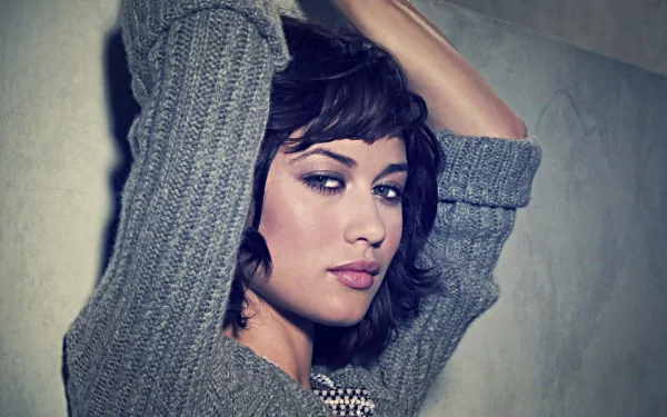HD PC desktop wallpaper featuring celebrity Olga Kurylenko in a cozy gray sweater, posing with arms raised against a muted background.