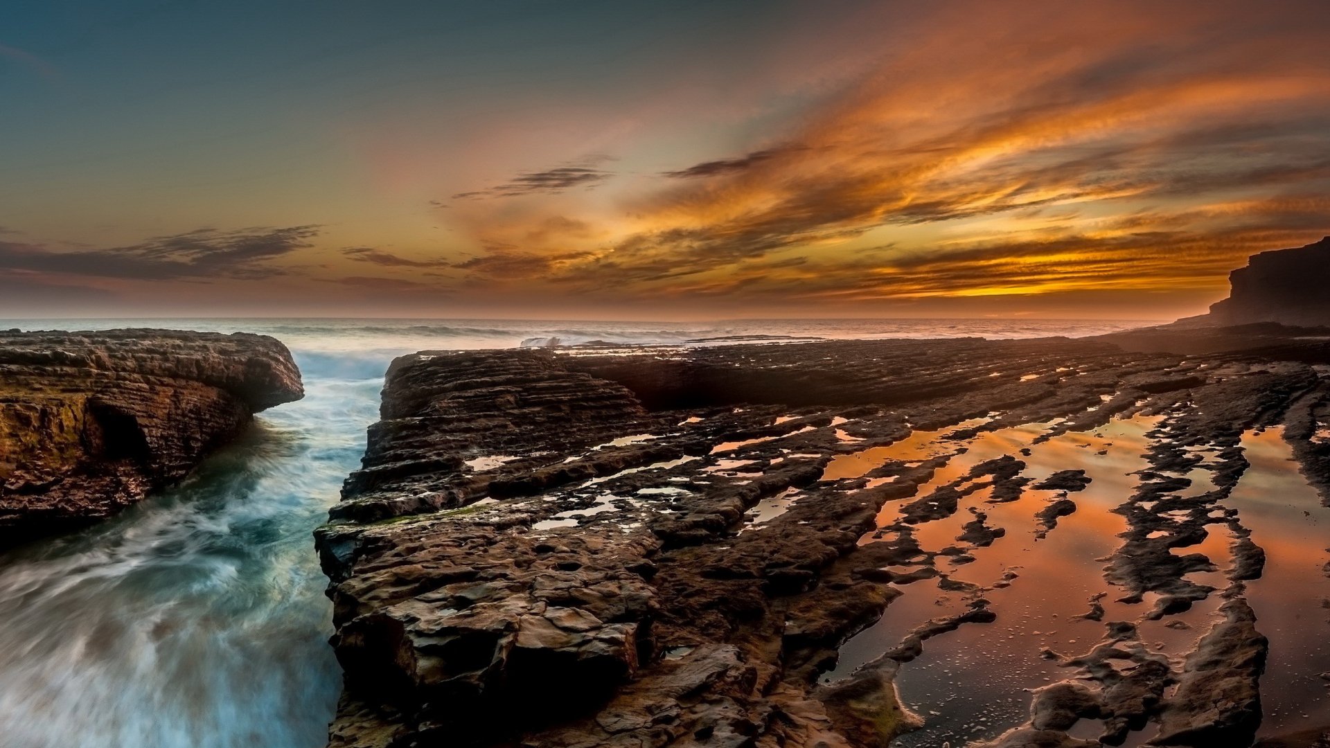 HD PC desktop wallpaper showing a dramatic ocean coastline at sunset with rocky cliffs and a vibrant sky reflecting on tidal pools.