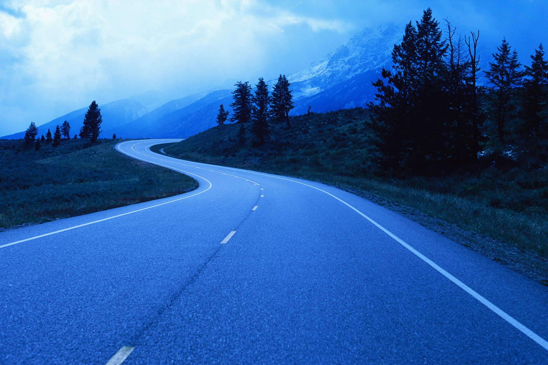 4K Ultra HD PC desktop wallpaper showing a man-made winding asphalt road curving through a blue-tinted mountain landscape with trees under a cloudy sky.