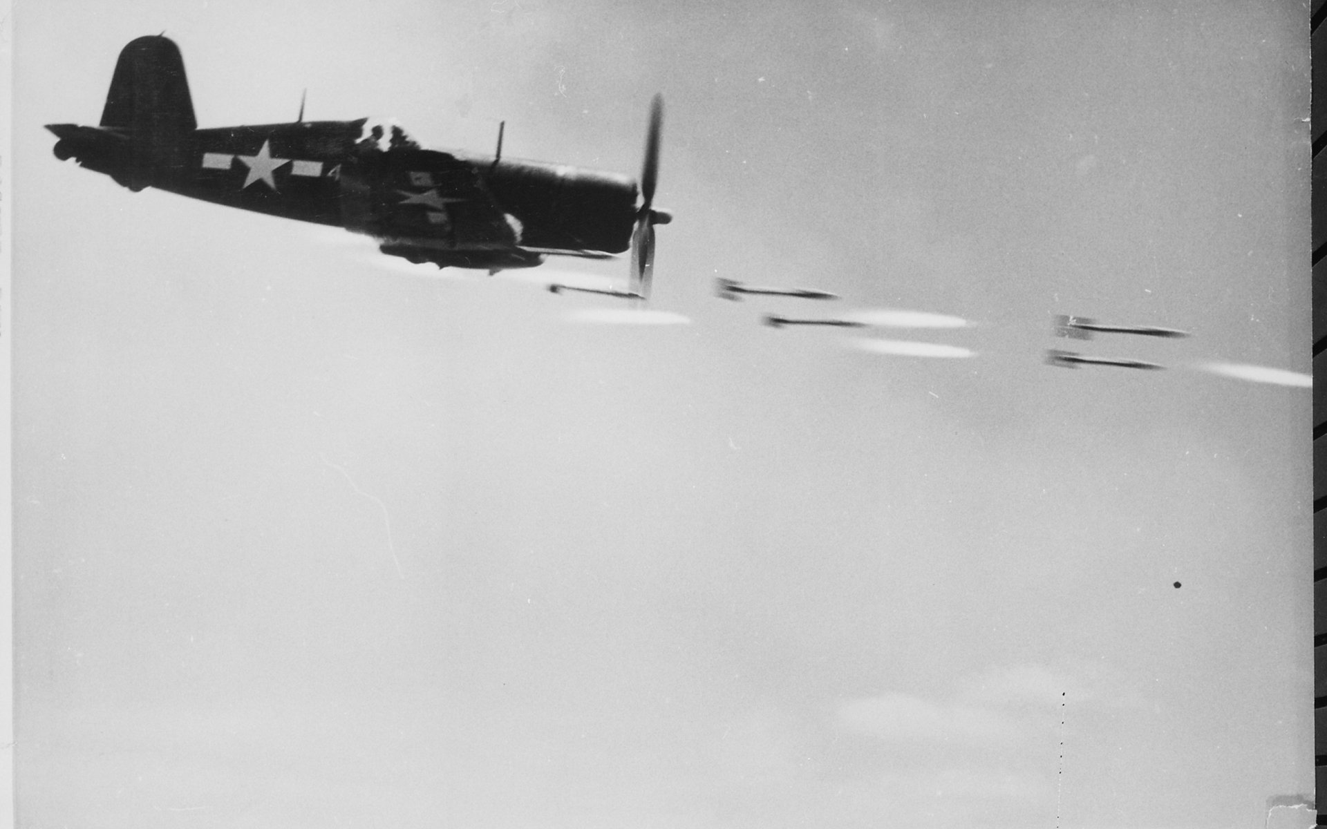 Black and white HD desktop wallpaper featuring a military Vought F4U Corsair fighter plane in flight, releasing rockets against a clear sky backdrop.