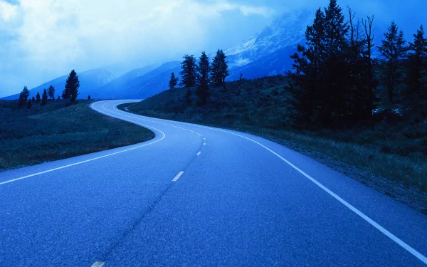 4K Ultra HD PC desktop wallpaper showing a man-made winding asphalt road curving through a blue-tinted mountain landscape with trees under a cloudy sky.