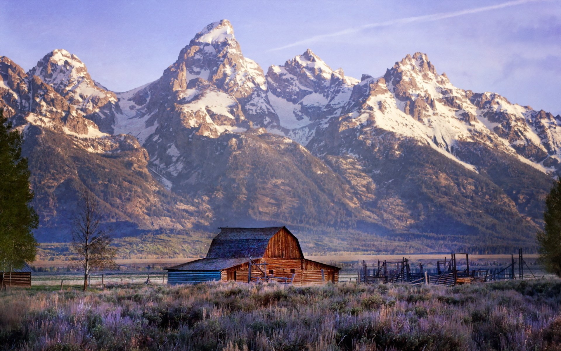 HD PC desktop wallpaper of a man-made wooden barn in a grassy field with snow-capped mountains rising behind under a pastel sky.