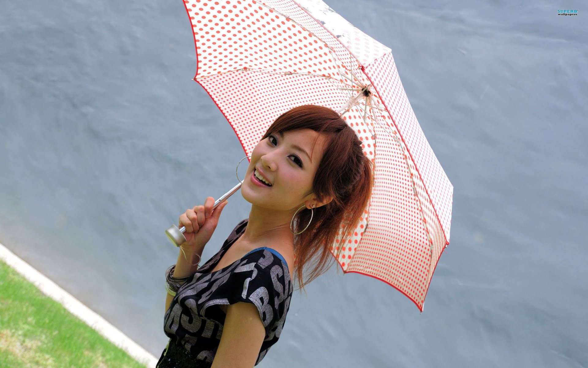 A woman, tagged as Mikako Zhang Kaijie, smiles while holding a red and white polka dot umbrella. She stands against a grayish background with a patch of green grass, in this HD desktop wallpaper.