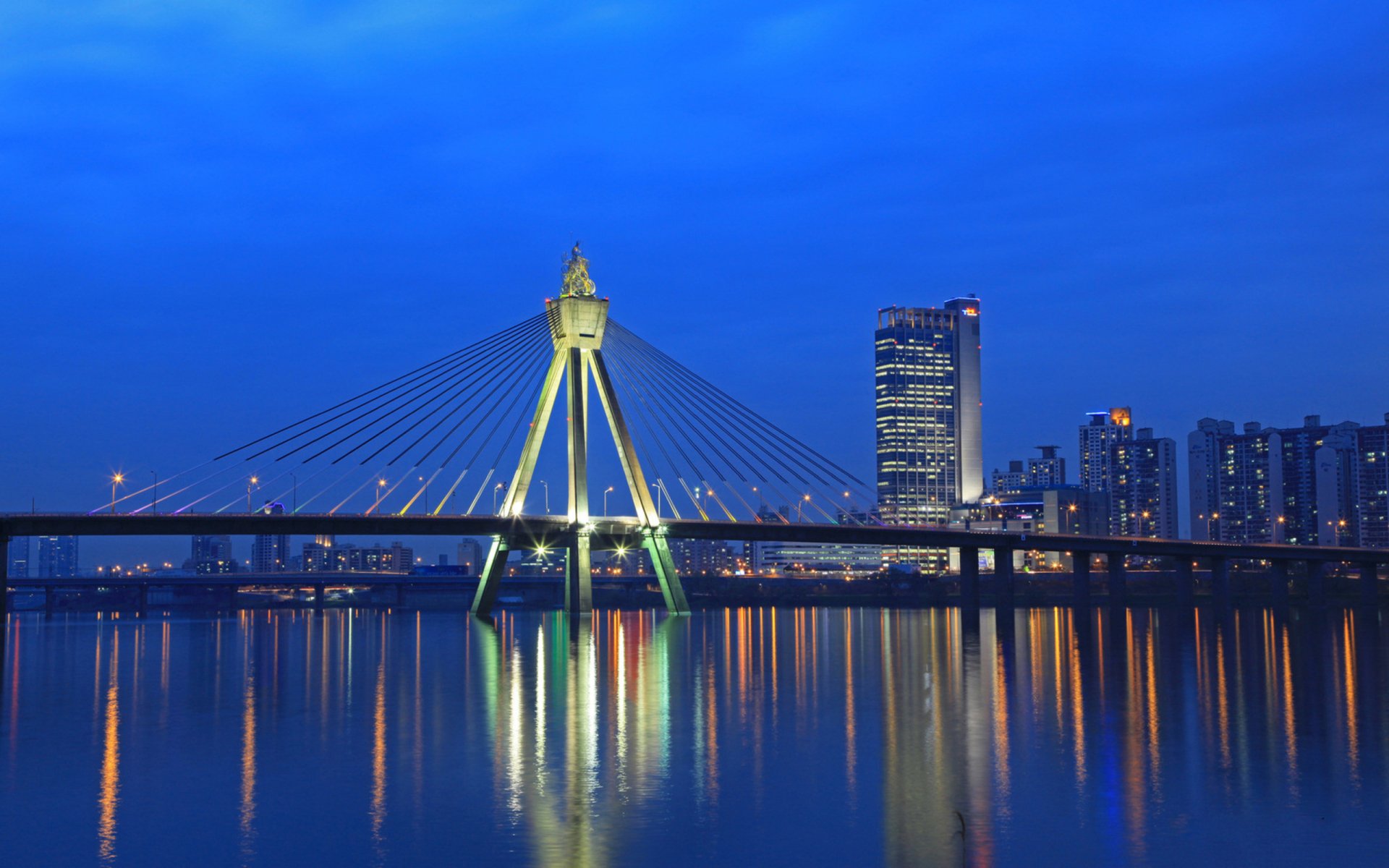 HD image of Seoul’s Olympic Bridge at night, showcasing its illuminated cables and reflection on the Han River with city buildings in the background.
