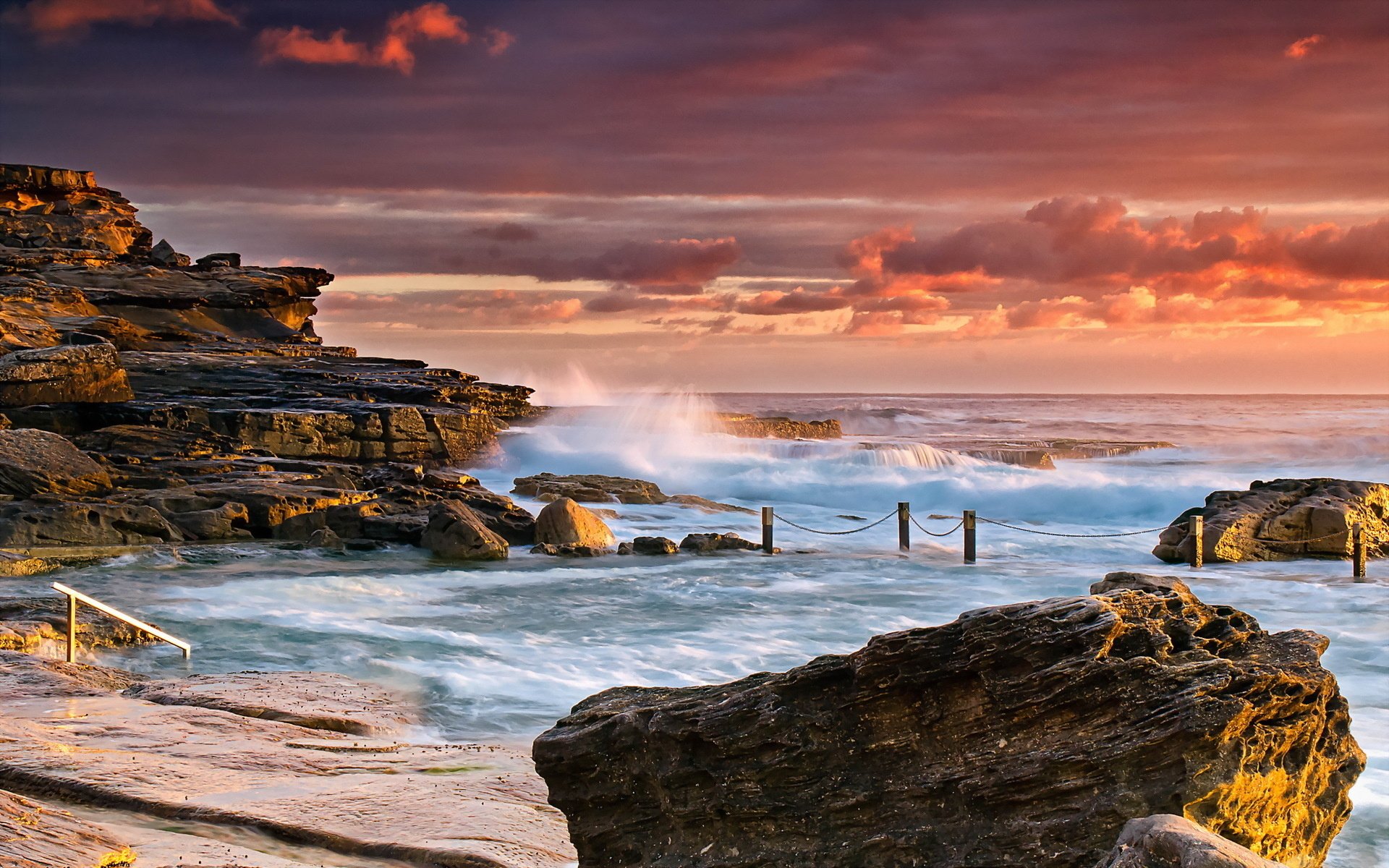 HD PC desktop wallpaper showing a rocky ocean shoreline at sunset with waves crashing and a colorful sky blending nature and sea elements.