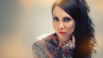 A captivating HD wallpaper featuring a woman with intricate tattoos, showcasing her striking features and intense gaze, set against a soft, blurred background.