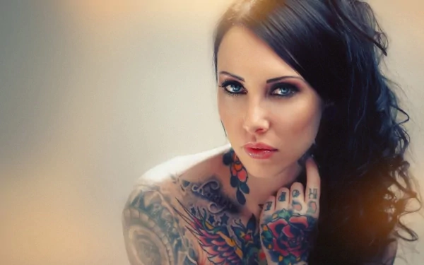 A captivating HD wallpaper featuring a woman with intricate tattoos, showcasing her striking features and intense gaze, set against a soft, blurred background.