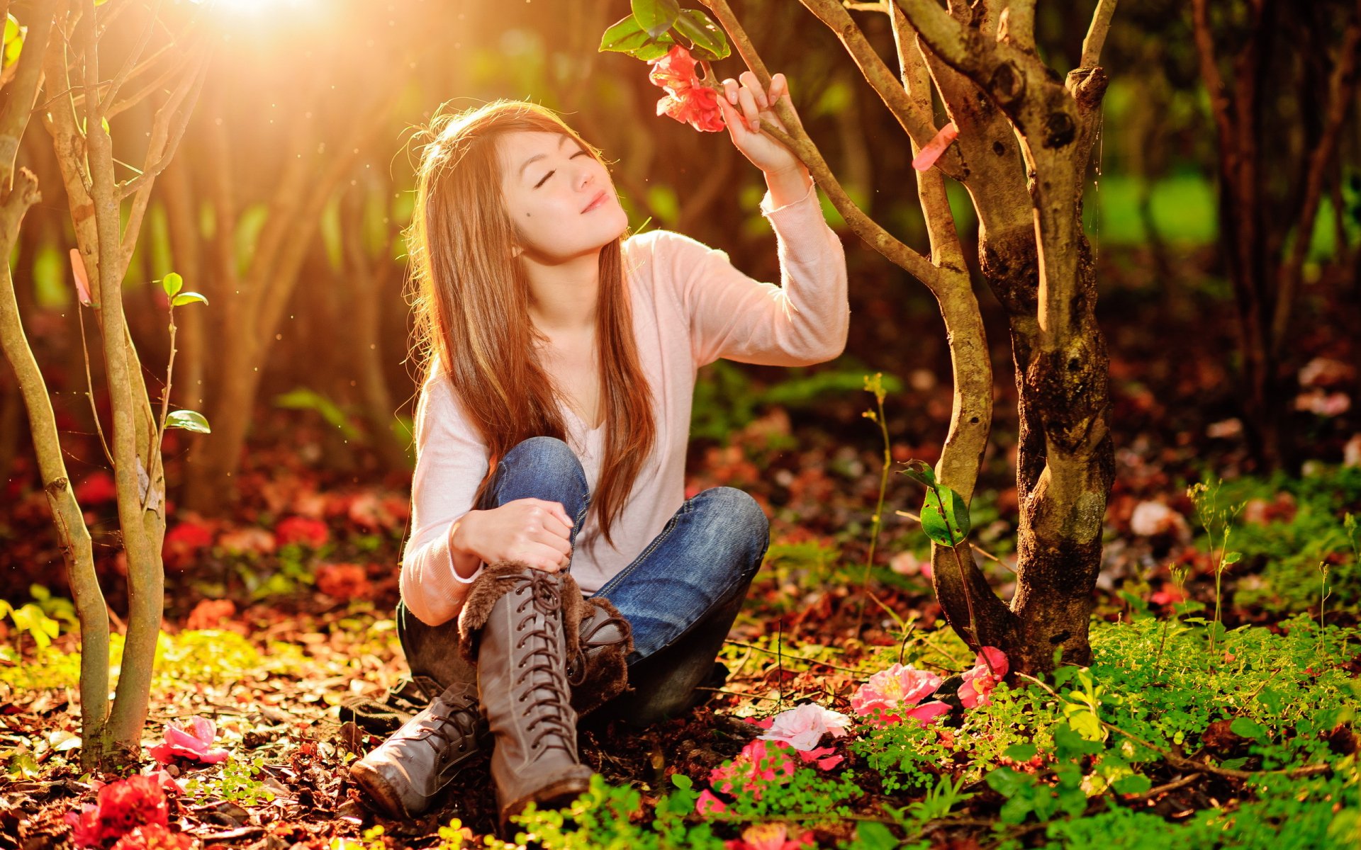An Asian woman sits peacefully on the ground among trees and colorful flowers, basking in sunlight. This HD desktop wallpaper captures a serene moment in nature.