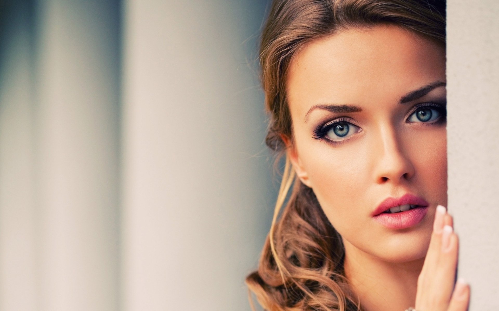 HD desktop wallpaper featuring a woman with blue eyes and blonde hair, looking directly into the camera while partially hidden behind a wall.