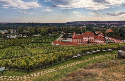 A panoramic HD desktop wallpaper of a man-made botanic garden in Prague, showcasing lush greenery, manicured paths, and historic buildings under a partly cloudy sky.