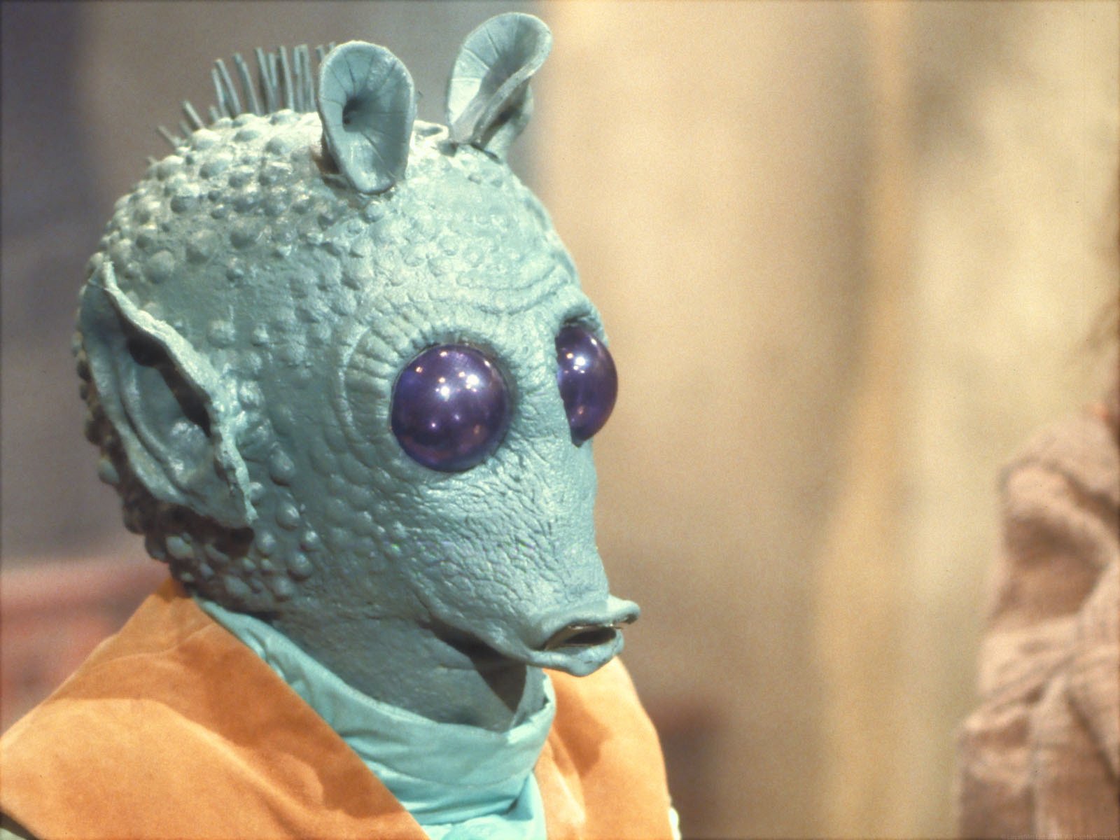 HD PC desktop wallpaper of Greedo — a green-skinned Star Wars Rodian with large purple eyes and spiky head, close-up portrait set against a softly blurred cantina background.