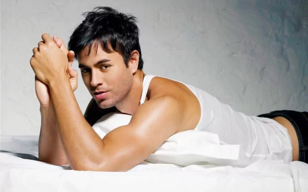 Enrique Iglesias poses casually on a bed, showcasing his muscular physique in a white tank top. This high-definition image serves as an engaging desktop wallpaper and background.