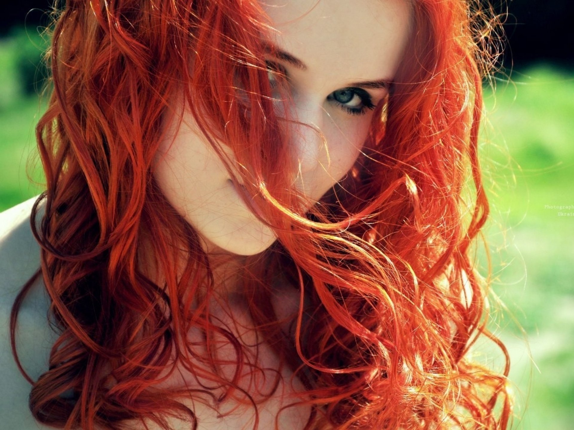 A close-up of a woman with vibrant red hair, her face partially obscured by flowing strands, set against a lush green background. This image serves as a striking HD desktop wallpaper.