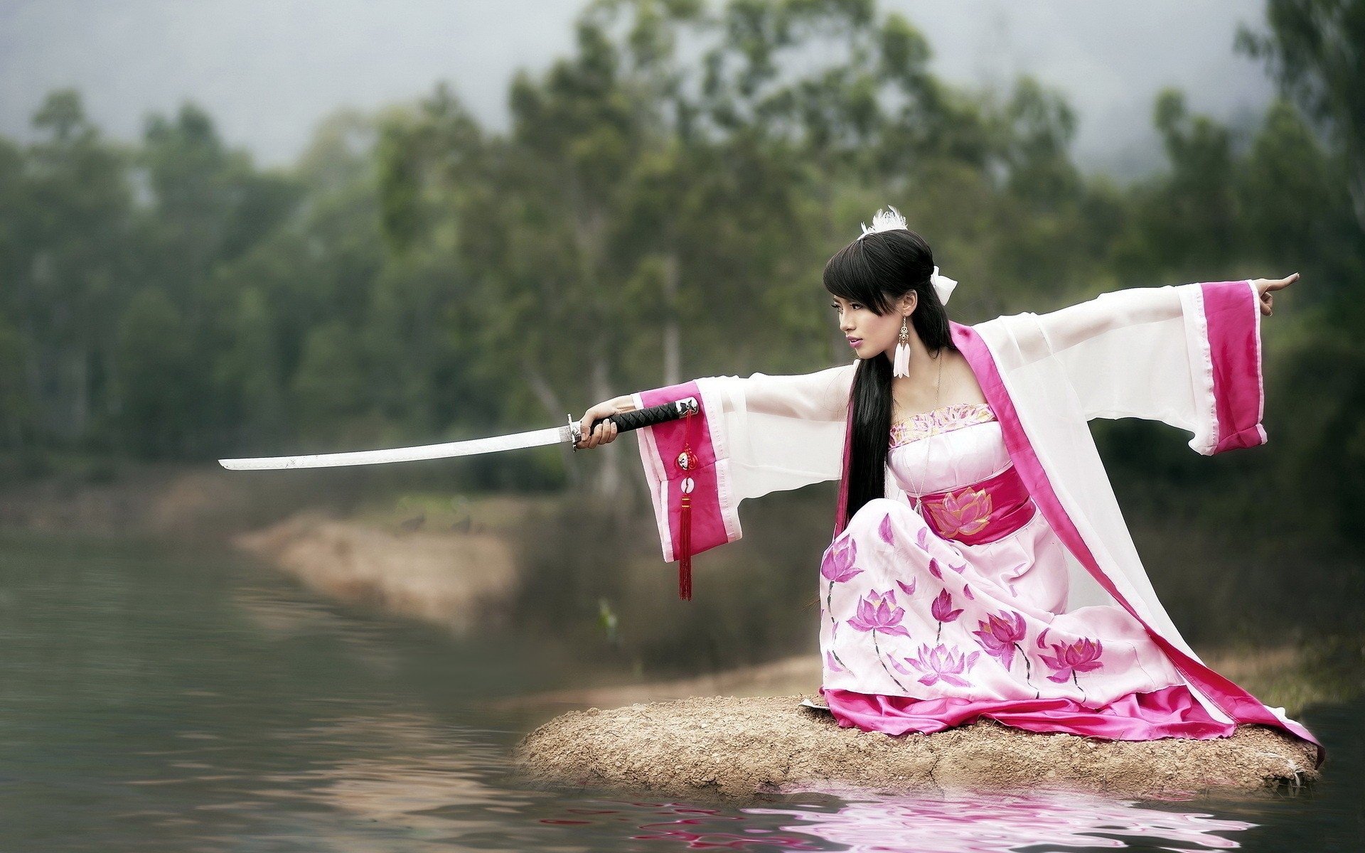 A woman in traditional attire stands in a serene landscape, holding a sword and poised gracefully over water. This HD wallpaper captures an elegant moment in nature.