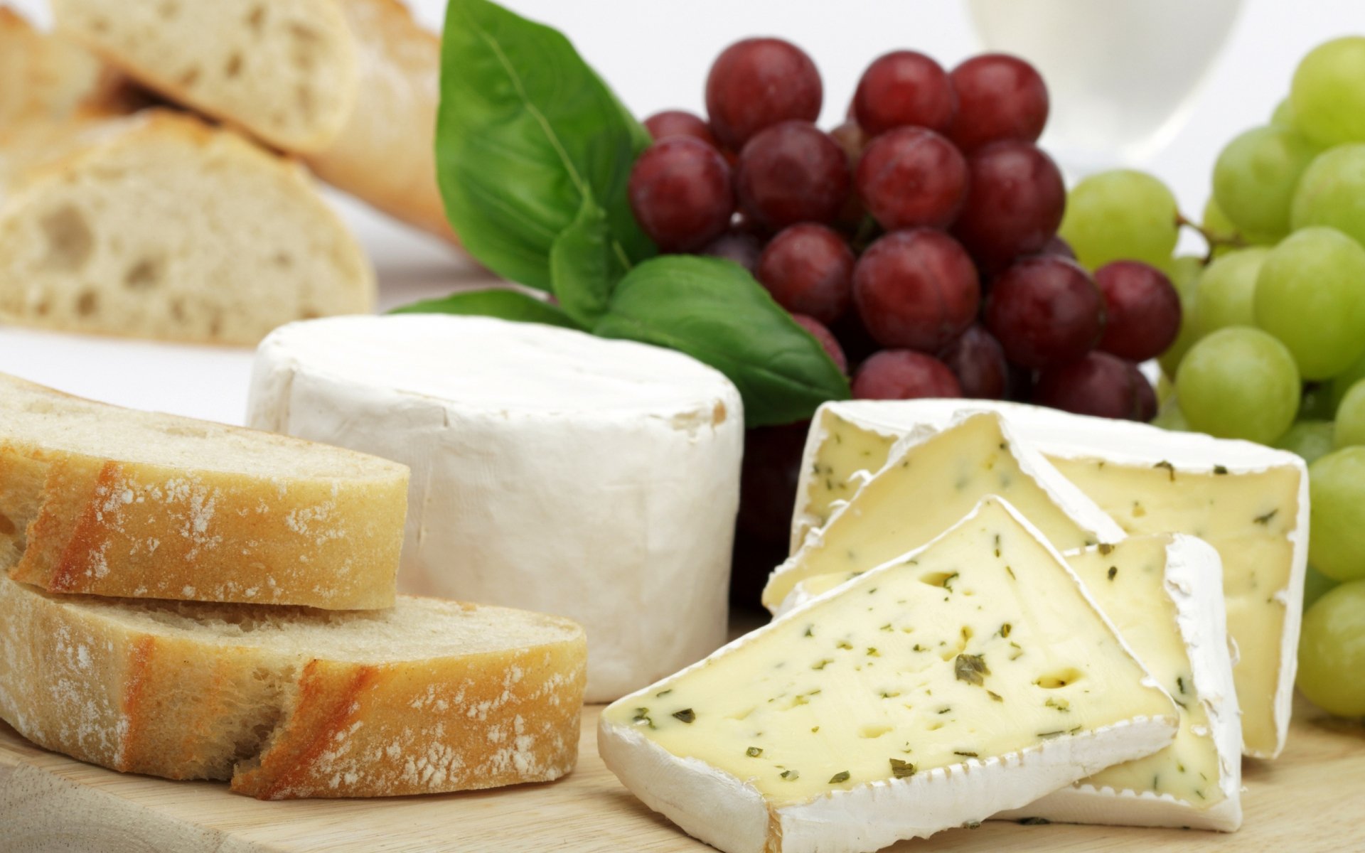 HD PC desktop wallpaper featuring a close-up of assorted cheeses, red and green grapes, fresh basil, and sliced bread arranged on a wooden board.