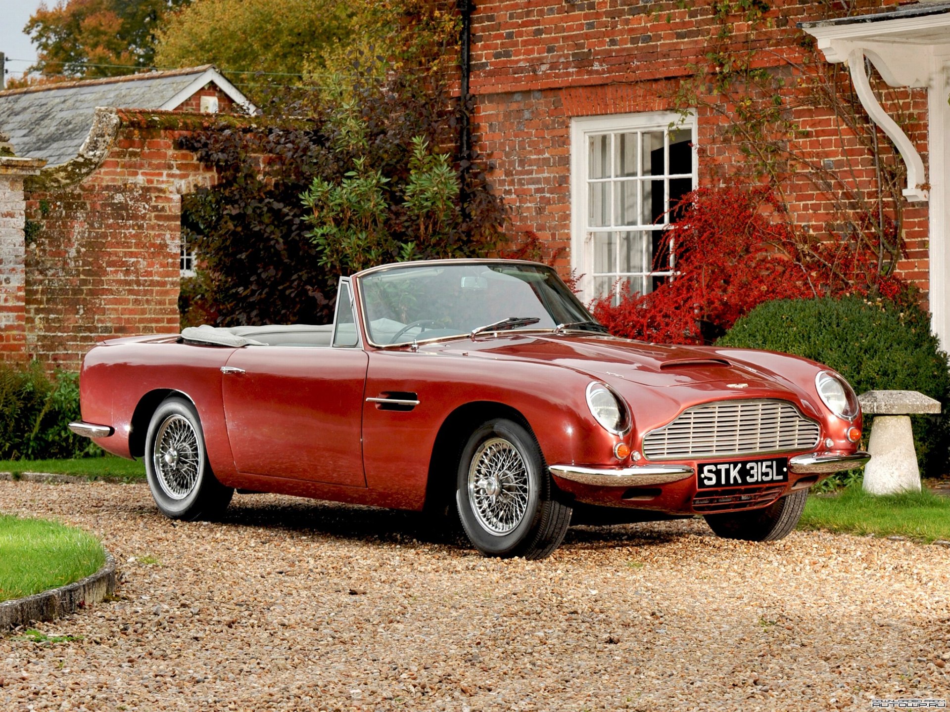HD PC desktop wallpaper featuring a classic red Aston Martin DB6 convertible parked on a gravel driveway in front of a rustic brick house with autumn foliage.