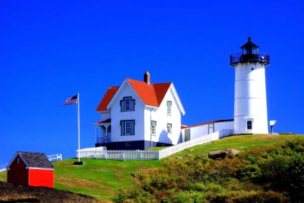 A charming lighthouse stands on a grassy hill in Martha's Vineyard, Massachusetts, accompanied by a quaint house and a small building, under a clear blue sky.