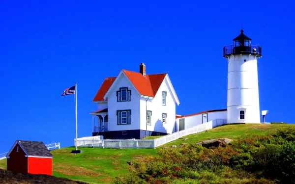 A charming lighthouse stands on a grassy hill in Martha's Vineyard, Massachusetts, accompanied by a quaint house and a small building, under a clear blue sky.