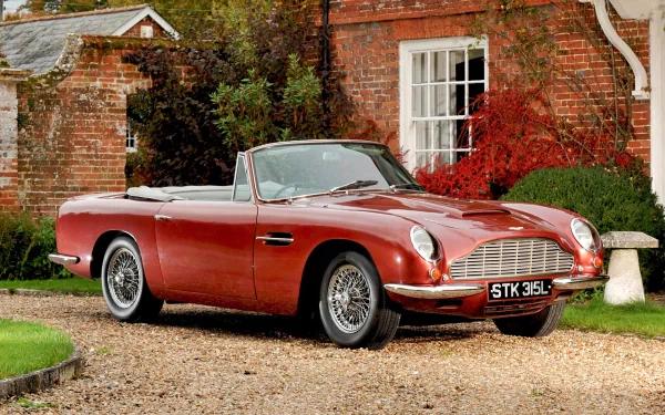 HD PC desktop wallpaper featuring a classic red Aston Martin DB6 convertible parked on a gravel driveway in front of a rustic brick house with autumn foliage.