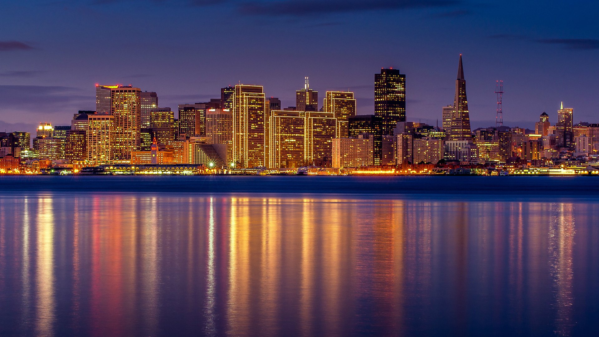 HD PC desktop wallpaper showing a man-made San Francisco skyline at dusk, glowing skyscrapers and the Transamerica Pyramid reflected in calm bay waters.