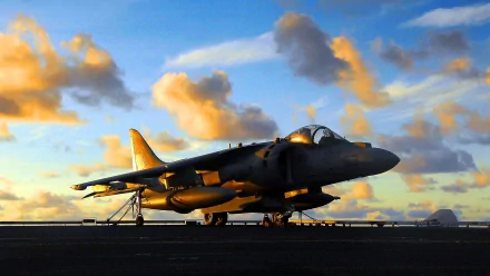 HD desktop wallpaper showing a McDonnell Douglas AV-8B Harrier II military jet on the runway during a vibrant sunset with scattered clouds.