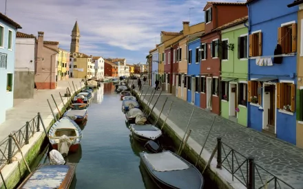 Colorful row houses along a Burano canal in Venice, Italy with moored boats and calm reflections under a blue sky — HD PC desktop wallpaper/background of a man-made town scene.