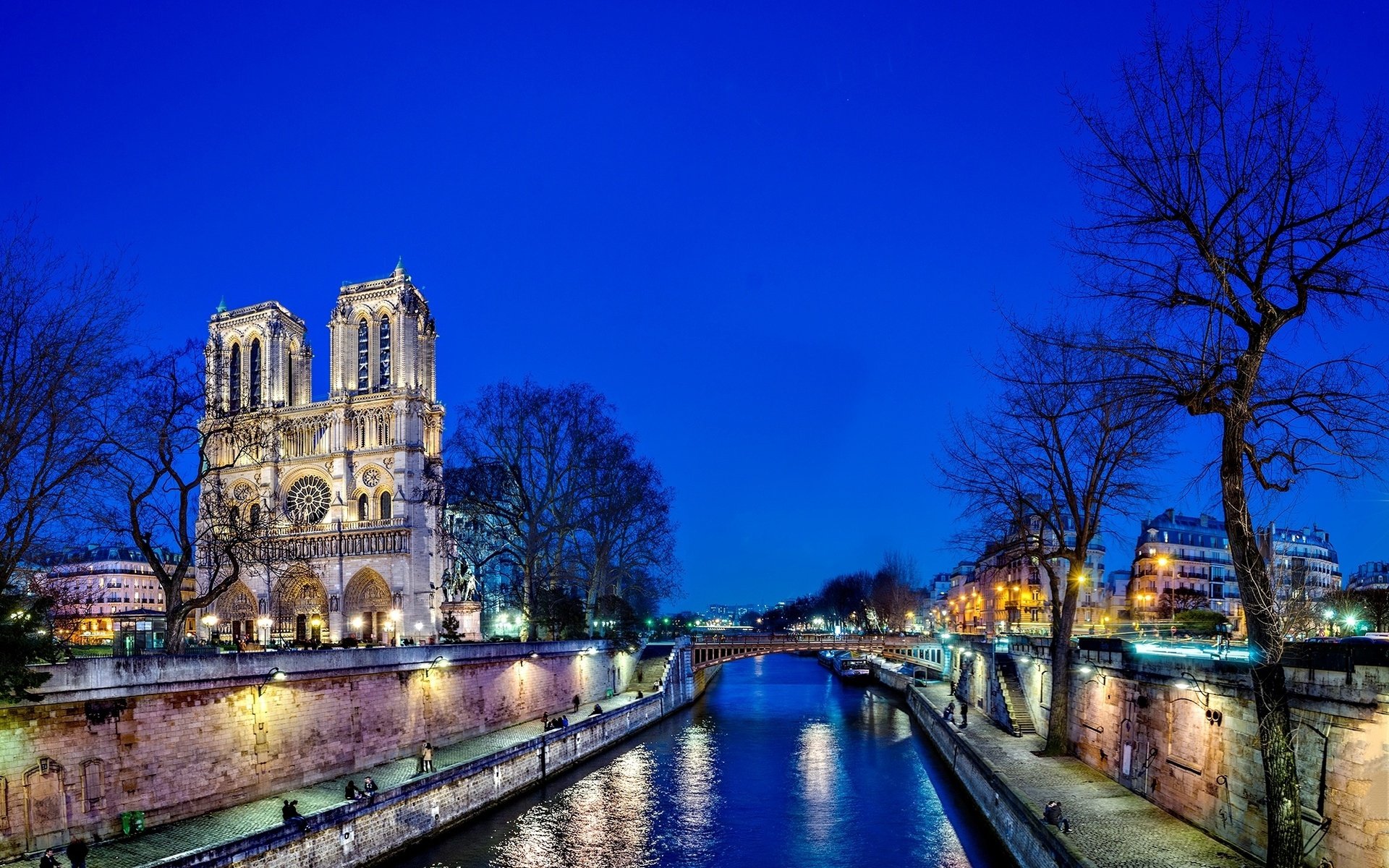 HD desktop wallpaper showcasing the illuminated Notre-Dame de Paris at night along the Seine River, highlighting its religious and historic grandeur.