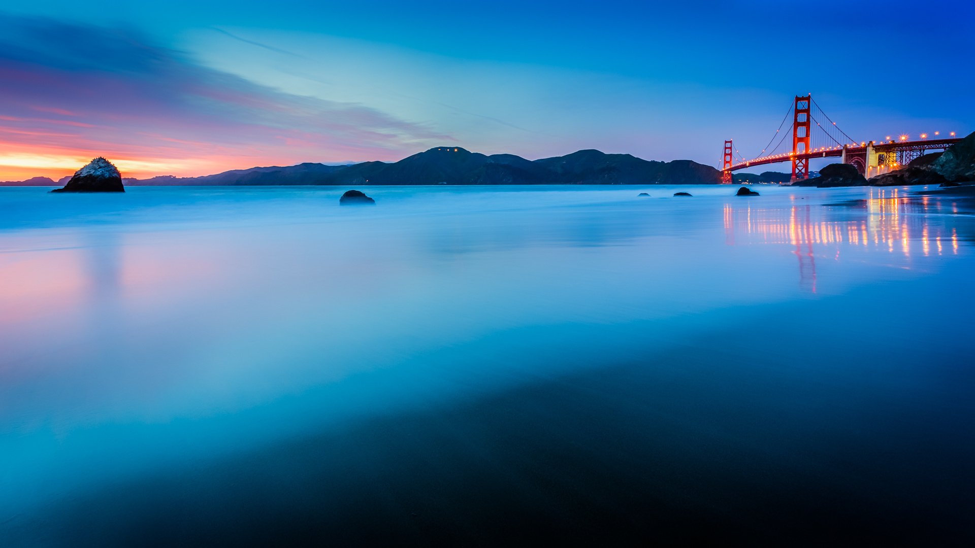 HD PC desktop wallpaper of the man-made Golden Gate Bridge at dusk, vibrant sunset sky reflected on a calm bay with silhouetted hills and the bridge's lights.