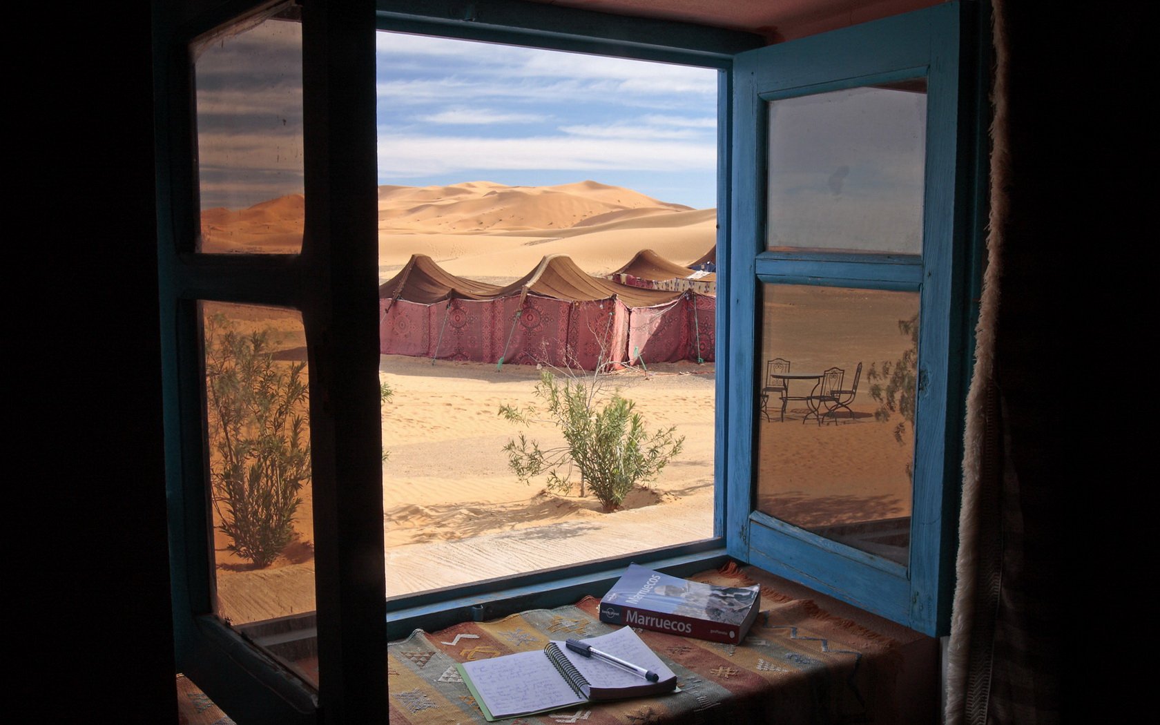 HD PC desktop wallpaper and background showing a man-made blue window framing desert tents and sand dunes; books and a notebook rest on the sunlit windowsill.