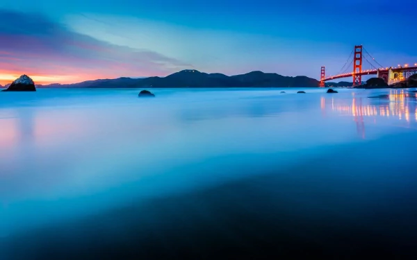 HD PC desktop wallpaper of the man-made Golden Gate Bridge at dusk, vibrant sunset sky reflected on a calm bay with silhouetted hills and the bridge's lights.