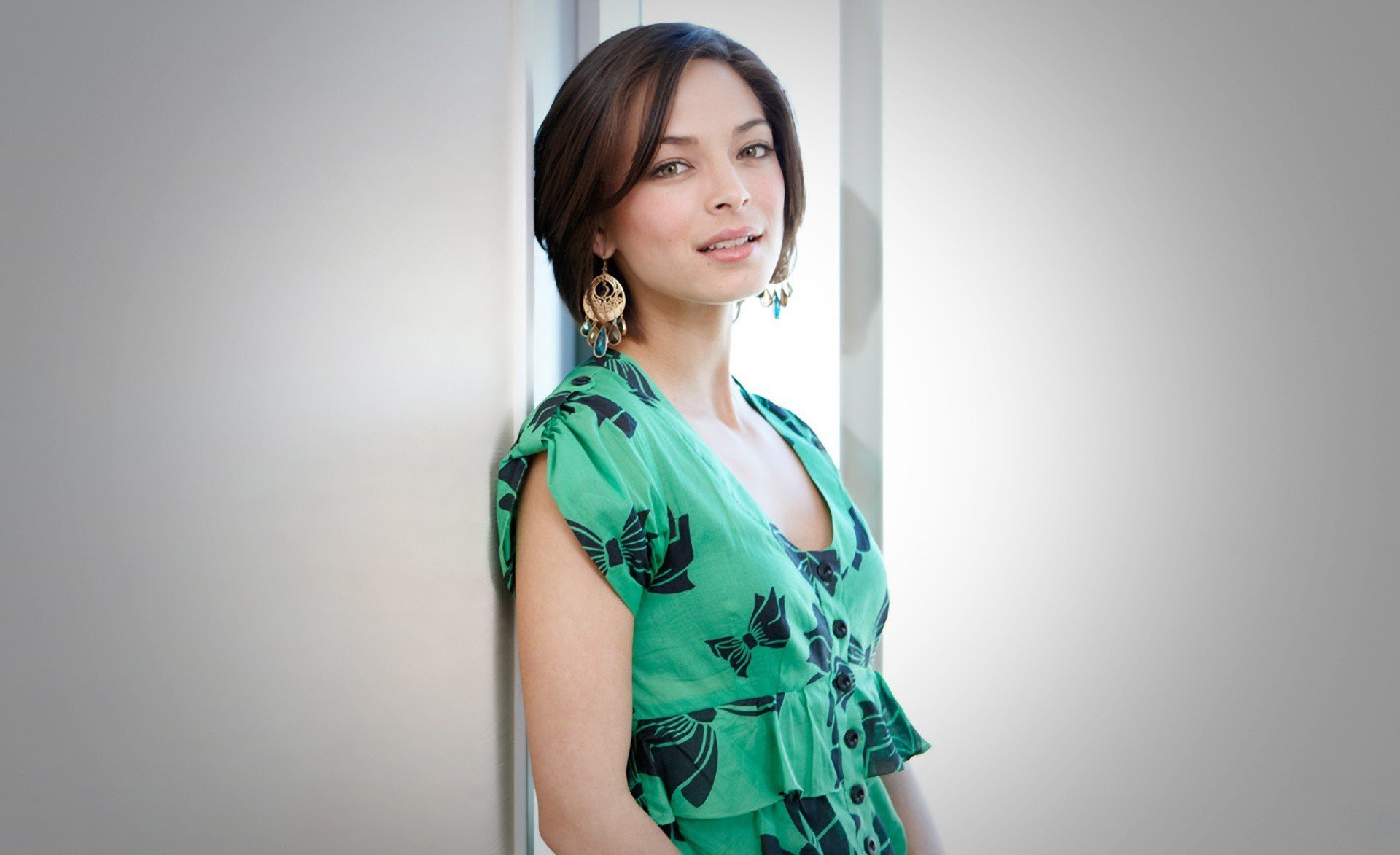 A high-definition desktop wallpaper featuring Kristin Kreuk, posed elegantly in a green outfit with floral patterns, showcasing her charm and style against a softly blurred background.