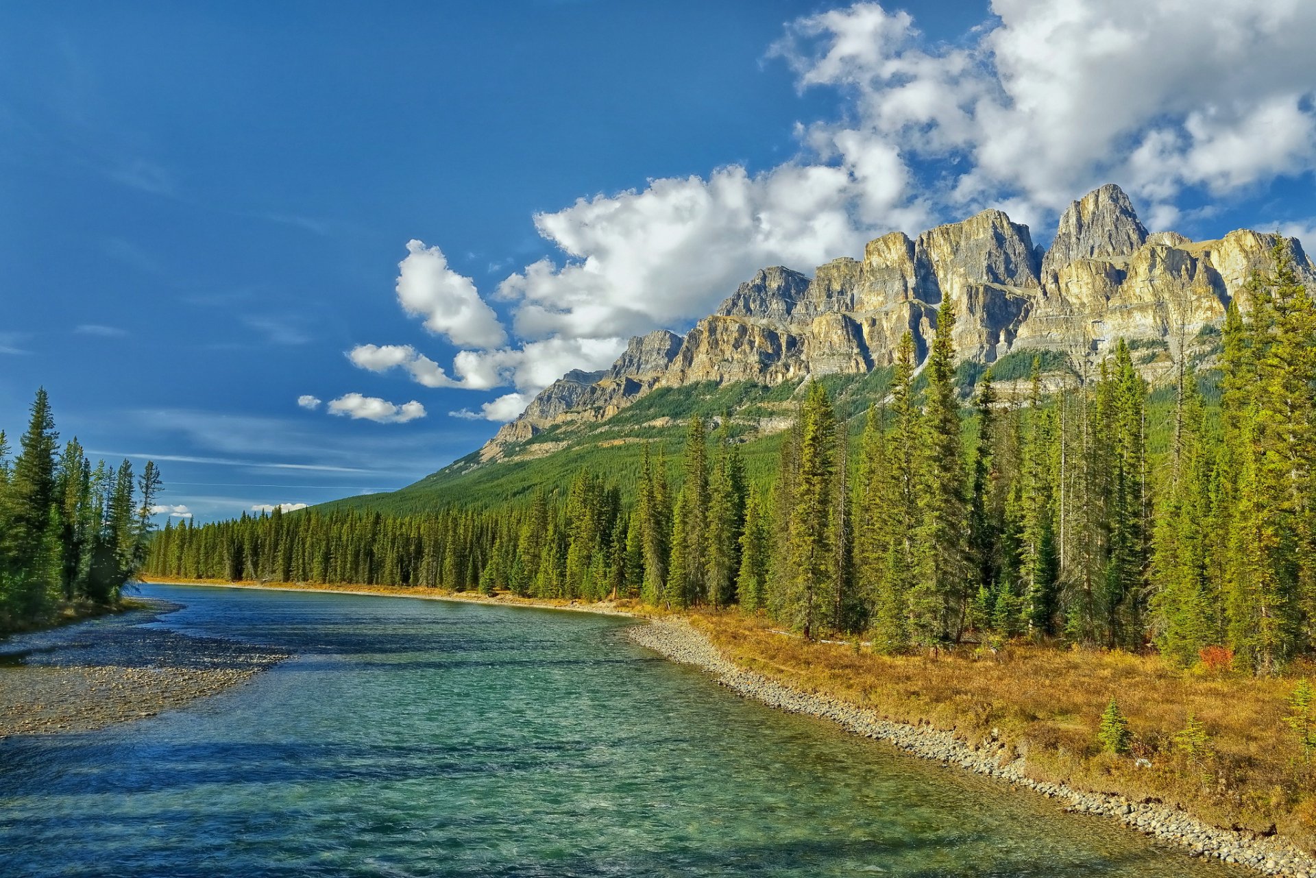 HD desktop wallpaper showcasing a clear river flowing beside a dense forest with towering mountains and a partly cloudy sky in the background.