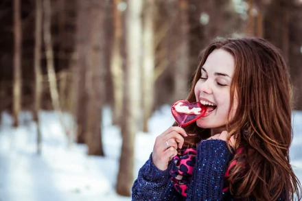 A woman enjoys a heart-shaped lollipop in a snowy forest, wearing a cozy sweater and colorful scarf. HD desktop wallpaper capturing a joyful, whimsical mood.