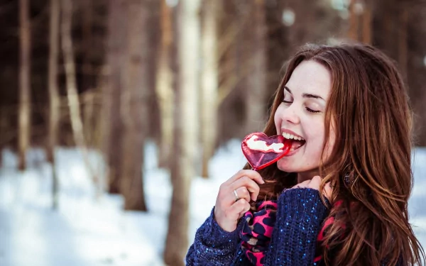 A woman enjoys a heart-shaped lollipop in a snowy forest, wearing a cozy sweater and colorful scarf. HD desktop wallpaper capturing a joyful, whimsical mood.