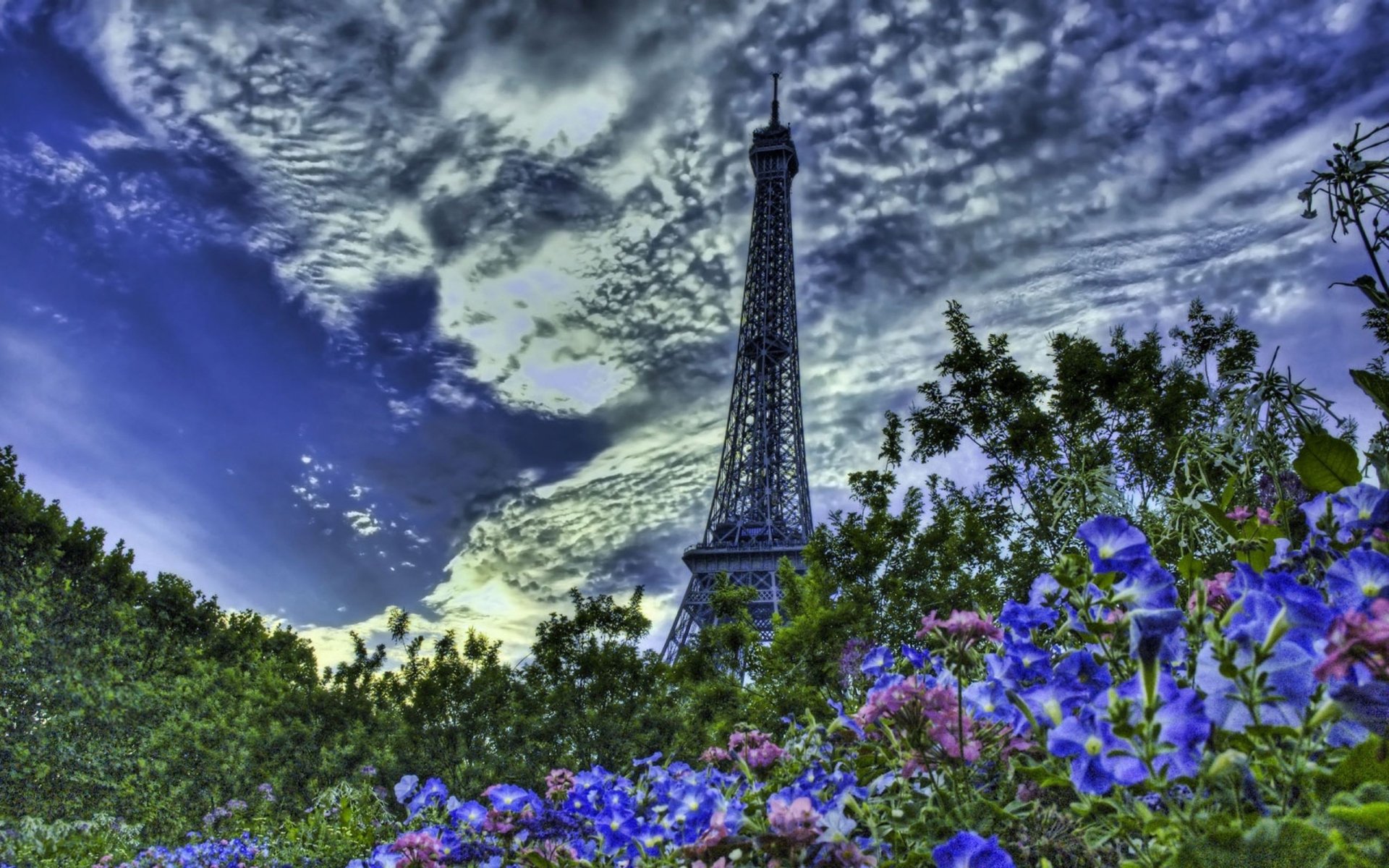 HD desktop wallpaper featuring the man-made Eiffel Tower towering above vibrant flowers and lush greenery under a dramatic, cloud-filled sky.