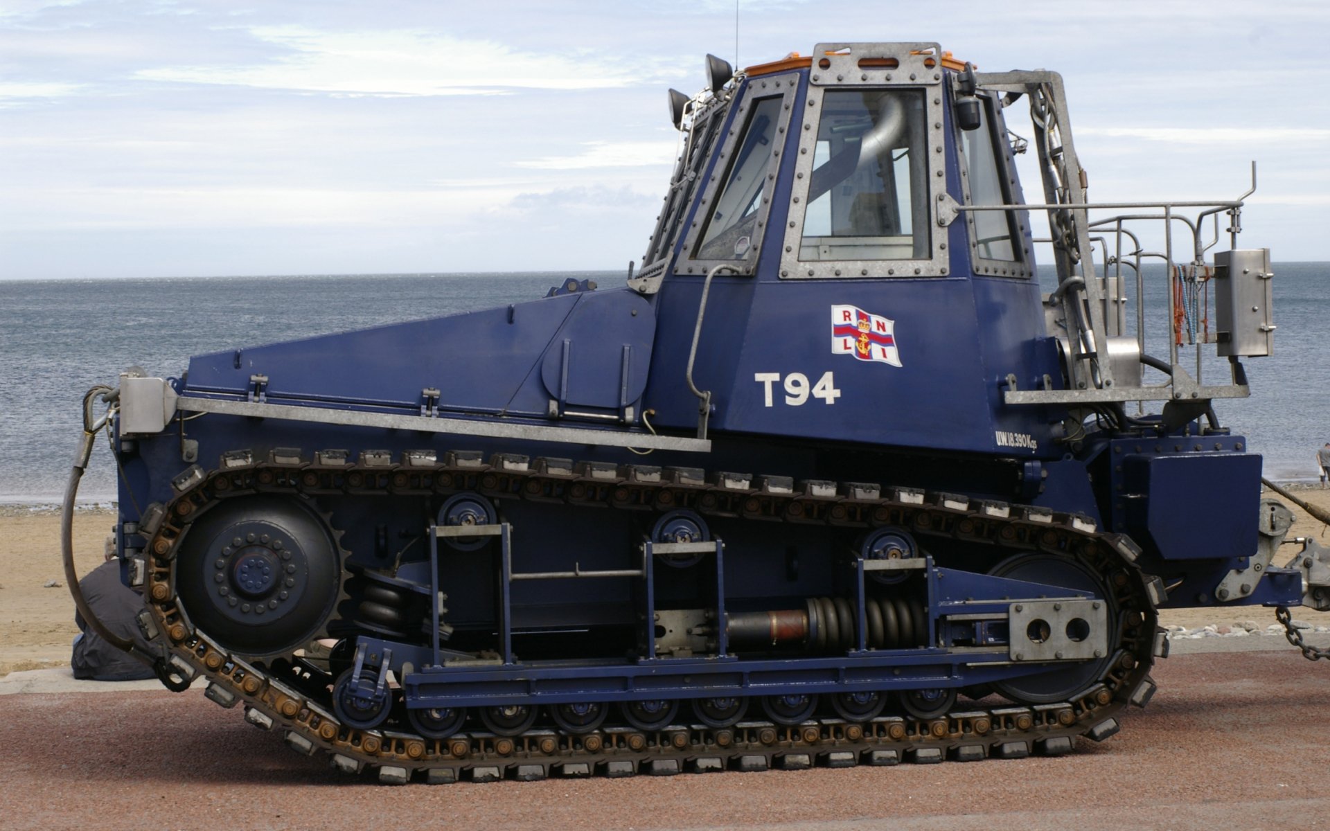 HD PC desktop wallpaper featuring a blue tracked tractor vehicle parked near a waterfront under a cloudy sky.