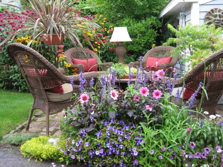 HD desktop wallpaper of a garden with wicker furniture, a lamp, and vibrant flowers. The scene depicts chairs and a table set amidst blooming plants, with a lush natural background.
