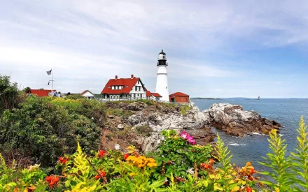 A serene lighthouse stands on rocky shorelines, surrounded by vibrant flowers and a picturesque house, creating a stunning HD desktop wallpaper and background.