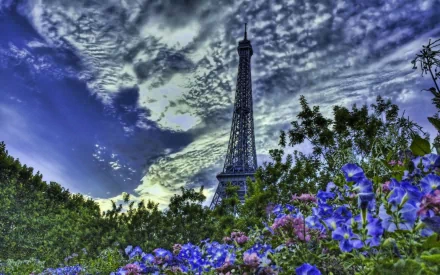 HD desktop wallpaper featuring the man-made Eiffel Tower towering above vibrant flowers and lush greenery under a dramatic, cloud-filled sky.
