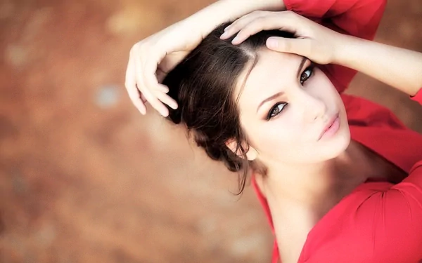 HD wallpaper featuring a beautiful woman with dark hair in a red outfit, gazing upwards while touching her head, set against a blurred background.