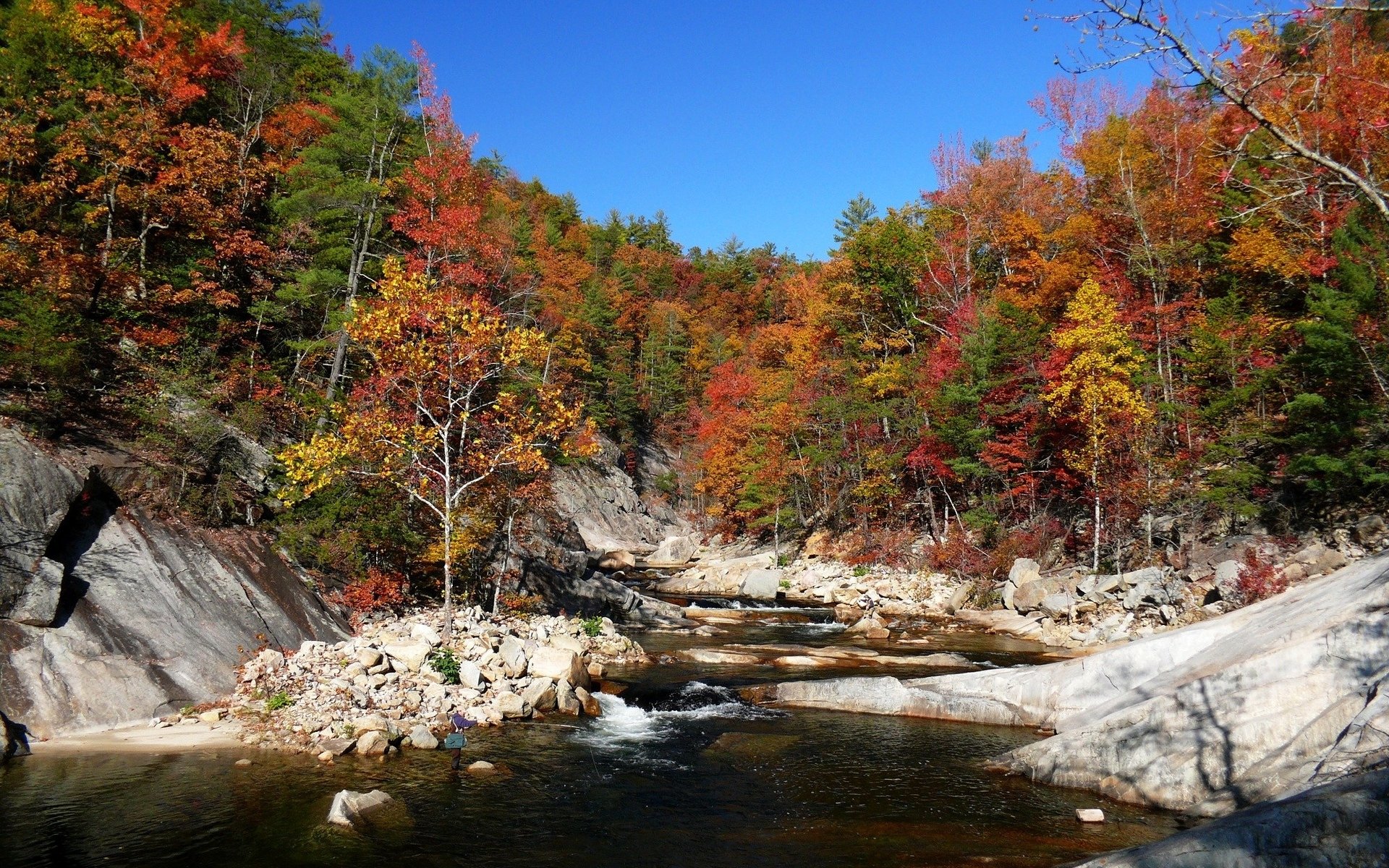 Vibrant fall foliage reflects on a serene river, surrounded by rocky banks and clear blue skies, captured in this stunning HD nature wallpaper.