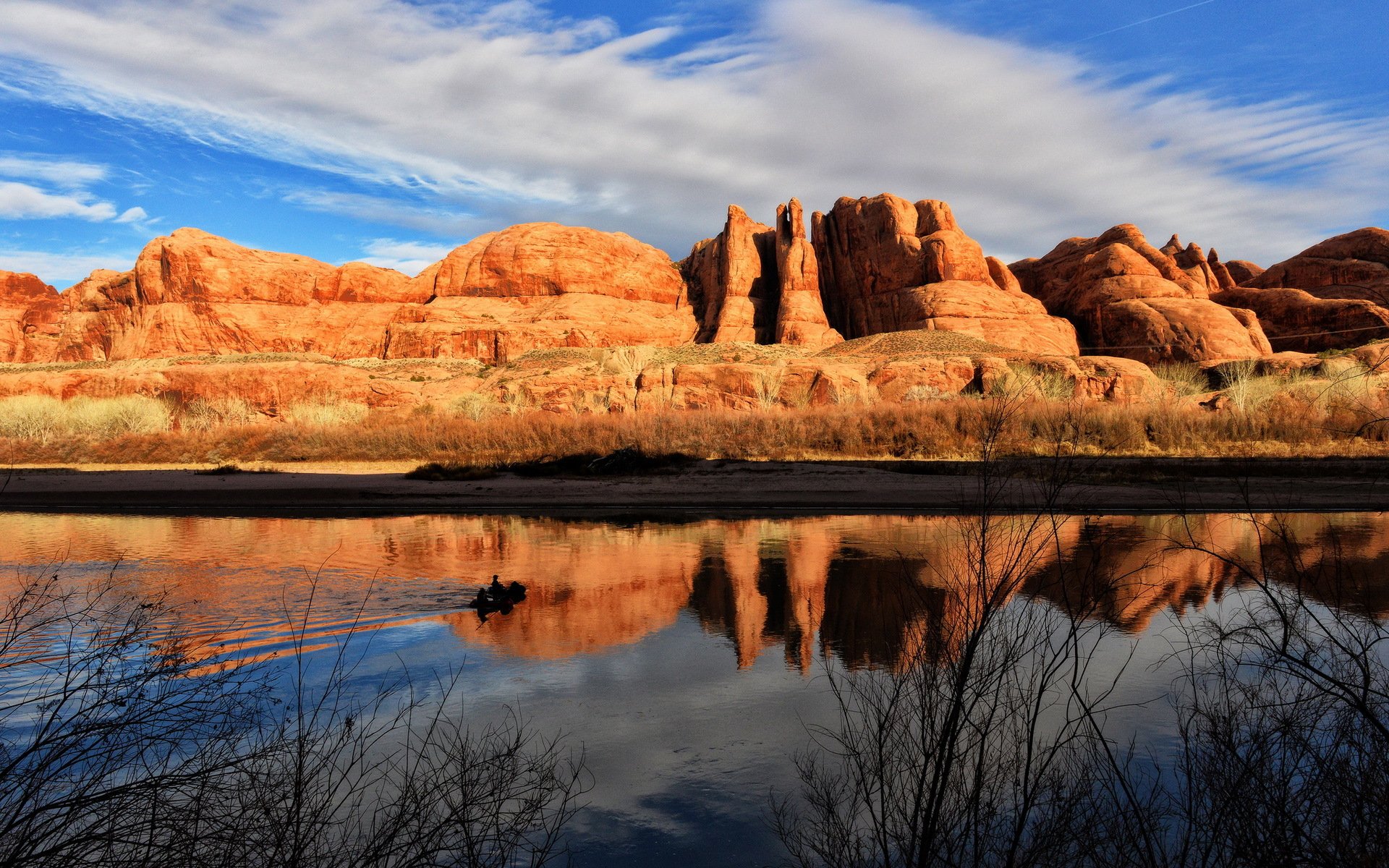 HD PC desktop wallpaper and background — desert nature scene of red sandstone cliffs under blue sky and clouds, mirrored in a calm river reflection with foreground shrubs.