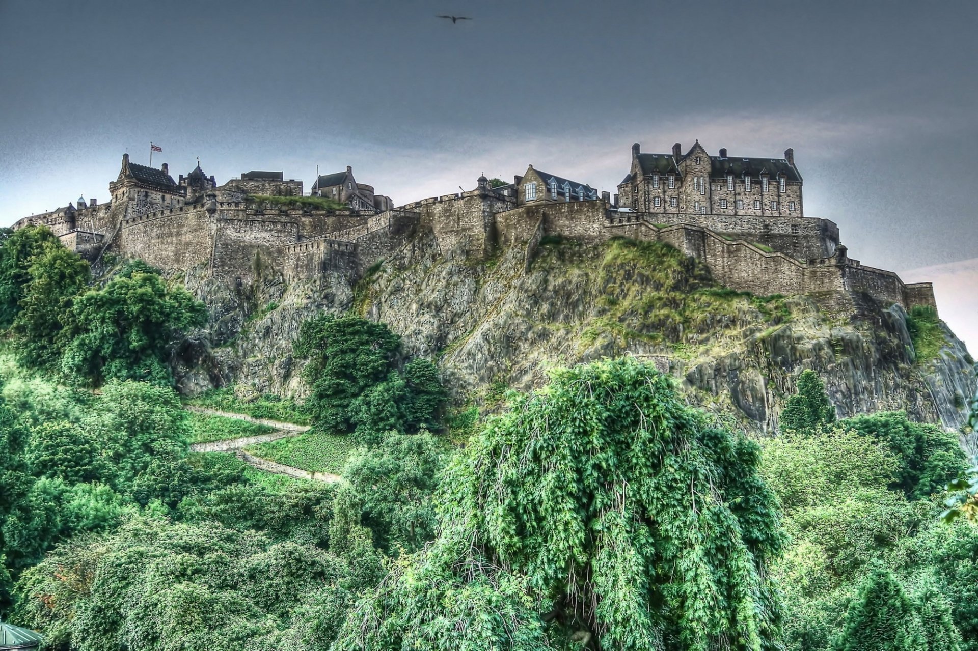 HD PC desktop wallpaper showcasing a majestic man-made castle perched atop a rocky hill, surrounded by lush green trees under a cloudy sky.