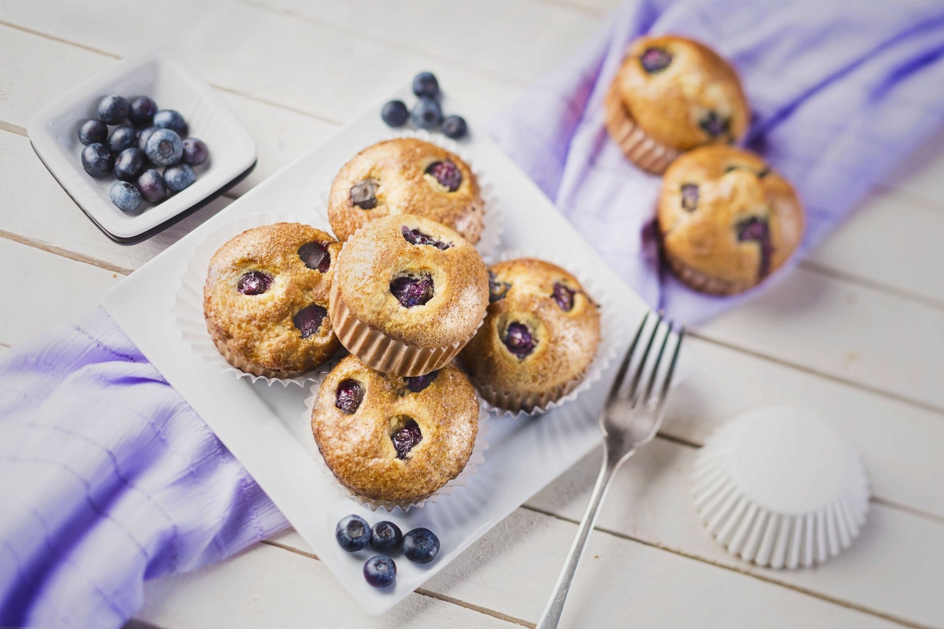 HD desktop wallpaper featuring freshly baked blueberry cupcakes on a white plate, with scattered blueberries and a soft purple cloth background.