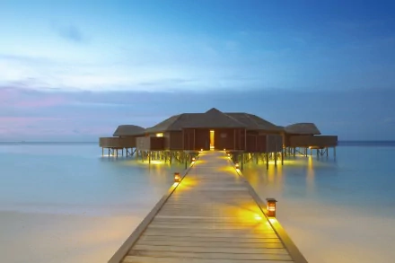 A serene ocean view features a wooden hut on stilts connected by a lit pier, basking in soft twilight hues. This captivating image makes an inviting HD desktop wallpaper.