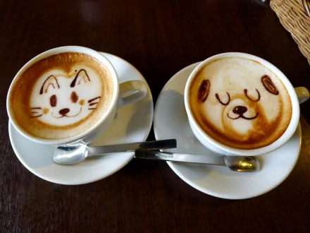 HD desktop wallpaper of two cups of coffee with cute latte art designs of a cat and a dog, placed on a dark wooden table.
